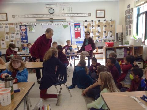 The Poet Laureate stands in a classroom addressing elementary-age students while a teacher looks on.