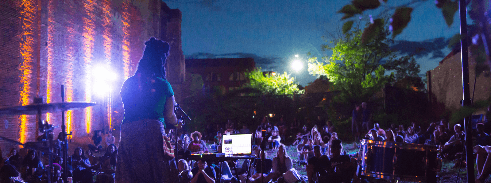 Person facing a seated outdoors crowd performing at night with back towards camera