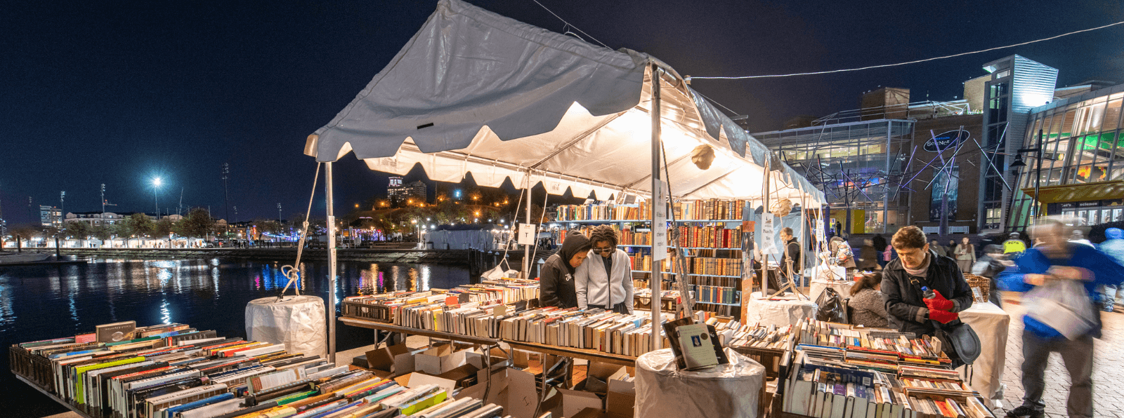 People browsing books at an outdoor tent next to a body of water