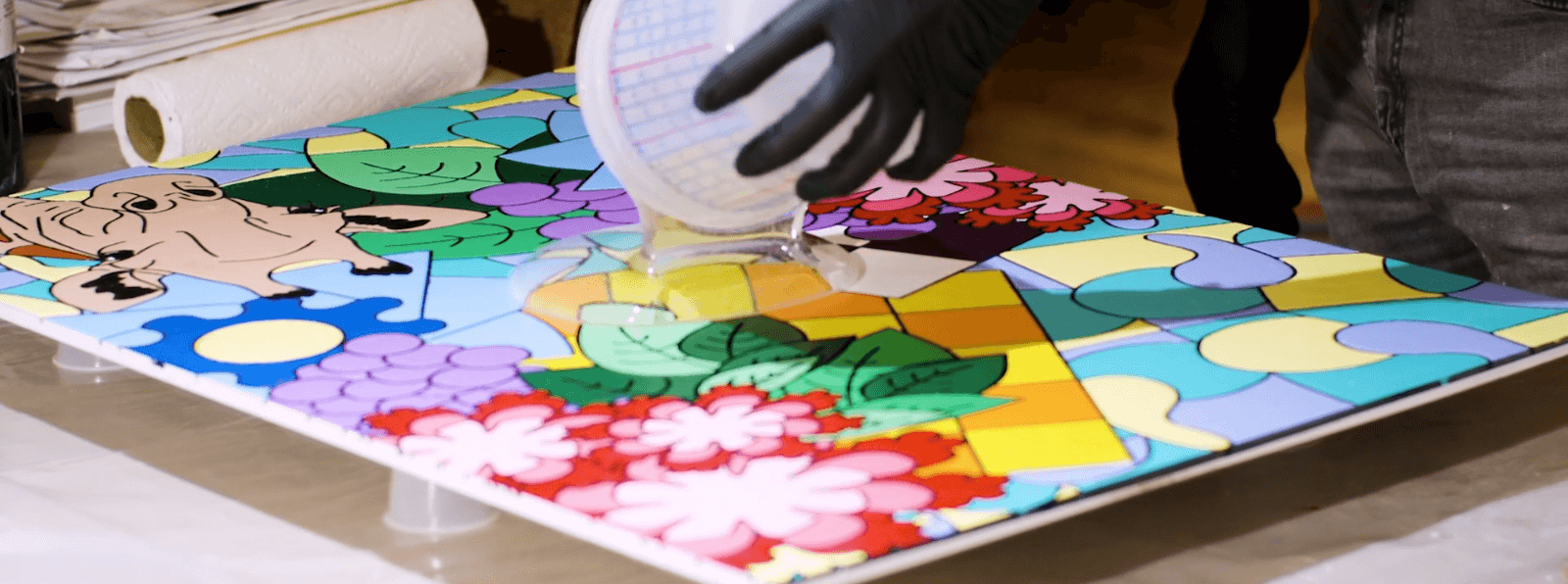 A person in gloves pouring clear liquid onto a red, green, yellow, orange, blue, and purple painting