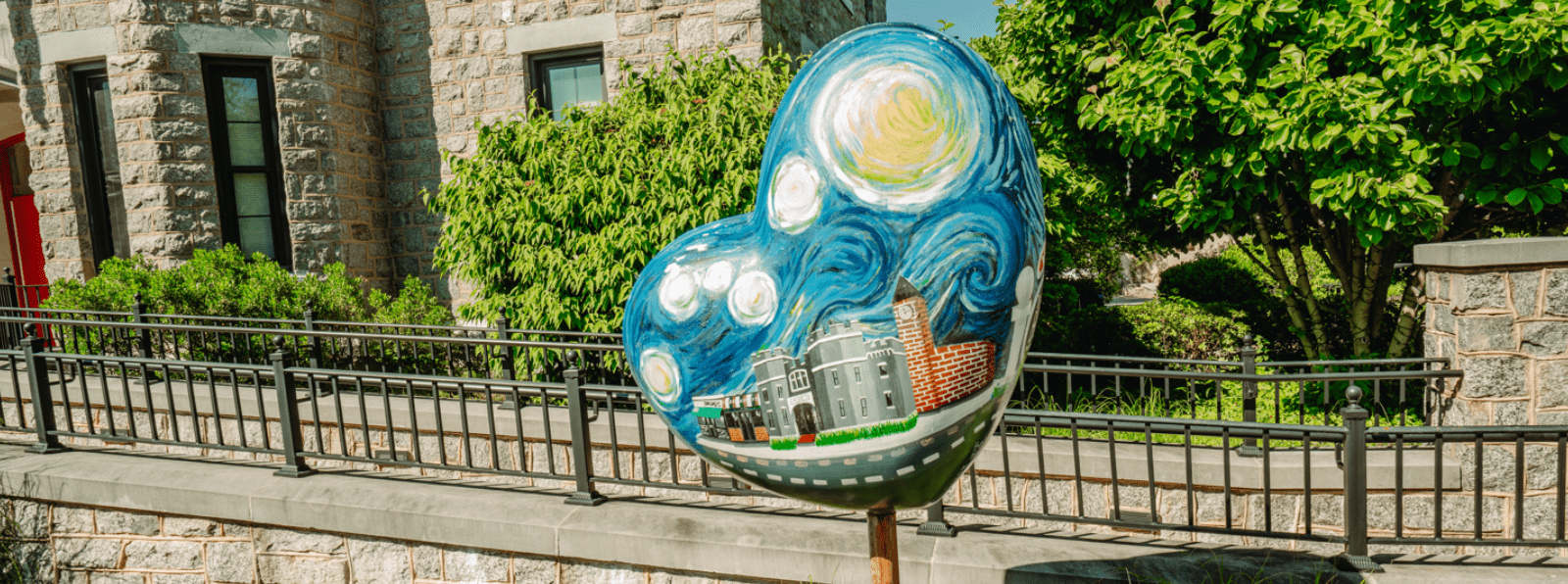 Heart shaped sculpture with a building and sky painted on it in front of a brick building