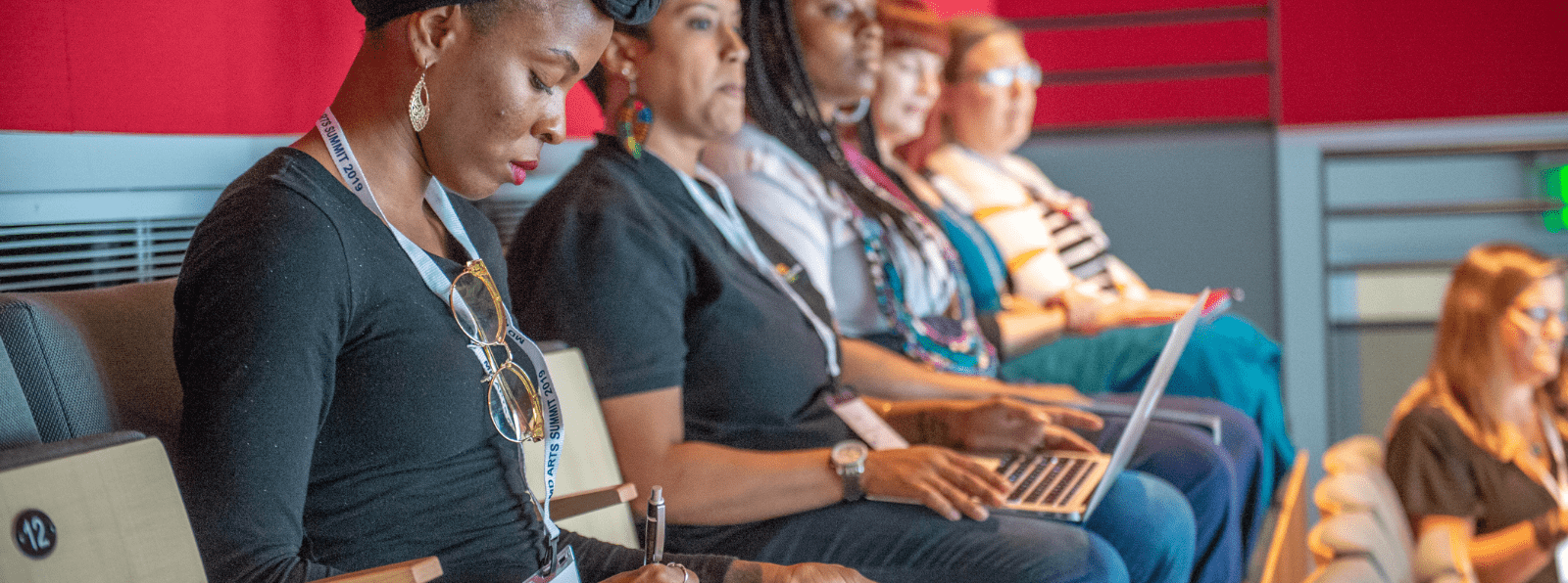 Five woman sit in a row taking notes by hand and on a laptop