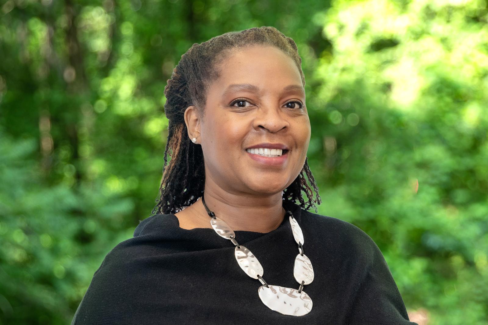 Monique N. Walker headshot. Woman smiling with white necklace and black shirt in front of green forest background.