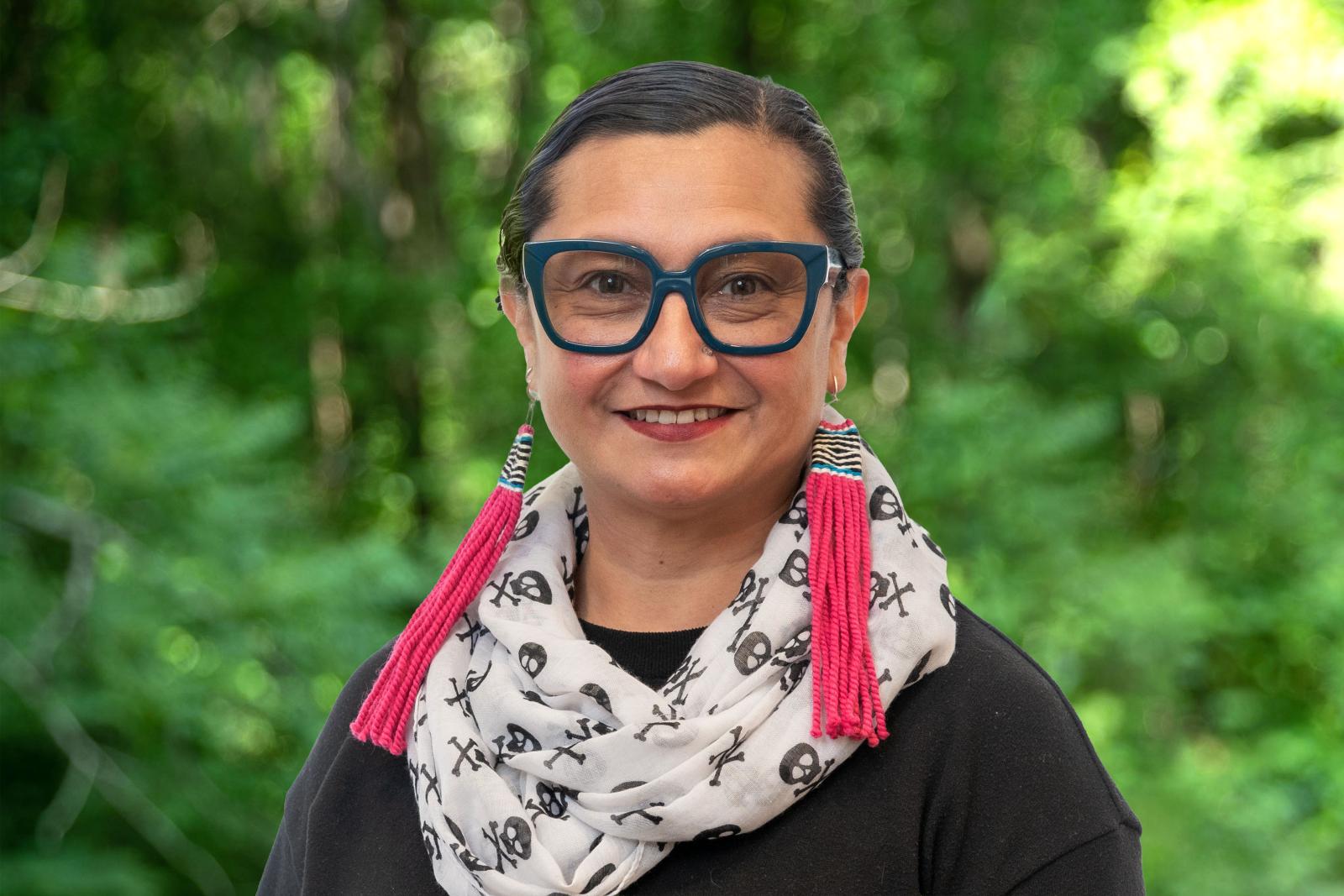 Headshot of Councilor Ruby Lopez Harper. Woman with glasses, white and black scarf and hot pink earrings in front of green forest background