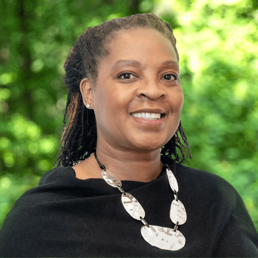 Monique N. Walker headshot. Woman smiling with white necklace and black shirt in front of green forest background.