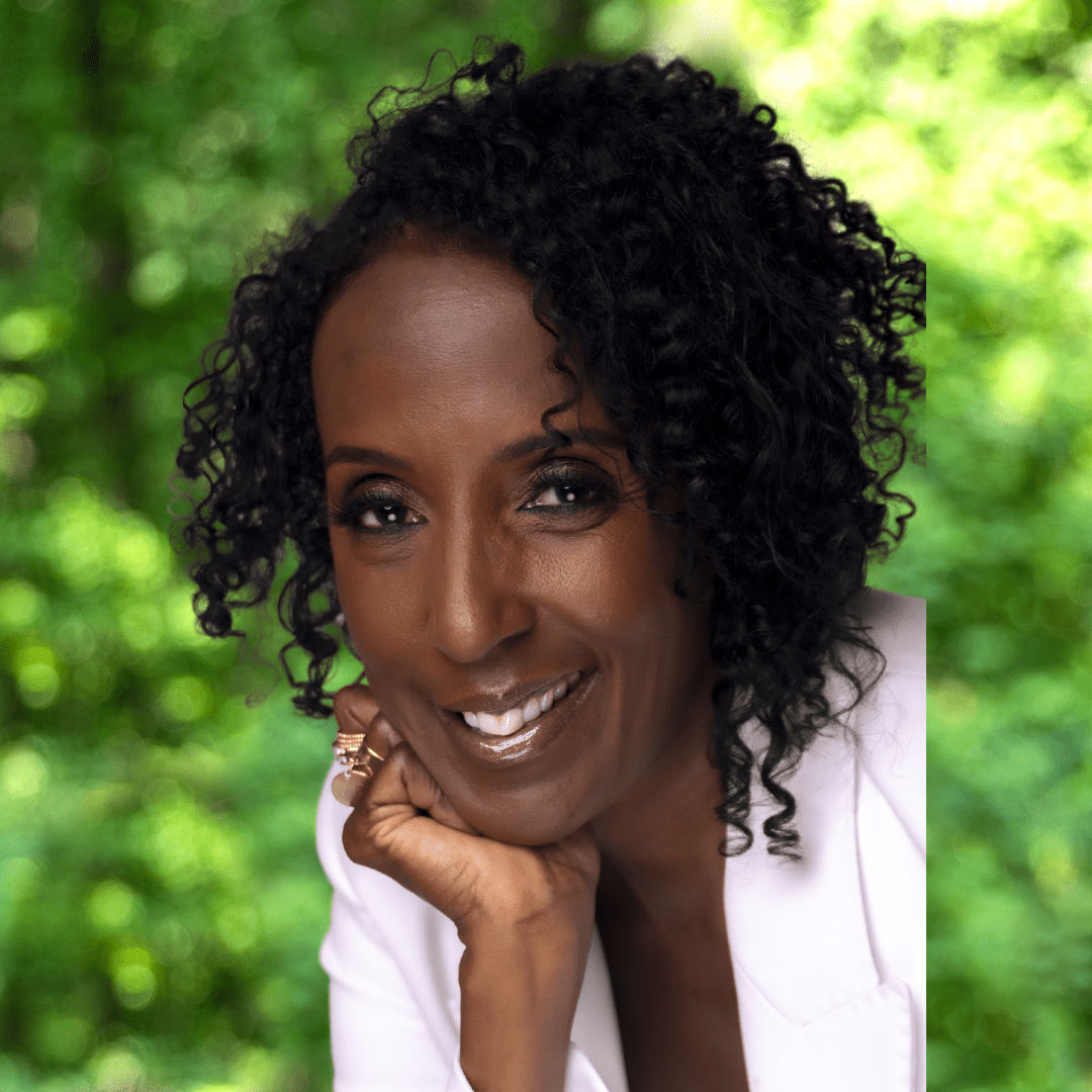 Smiling woman with curly black hair resting chin on closed hand, wearing a white suit jacket and looking directly at the camera.