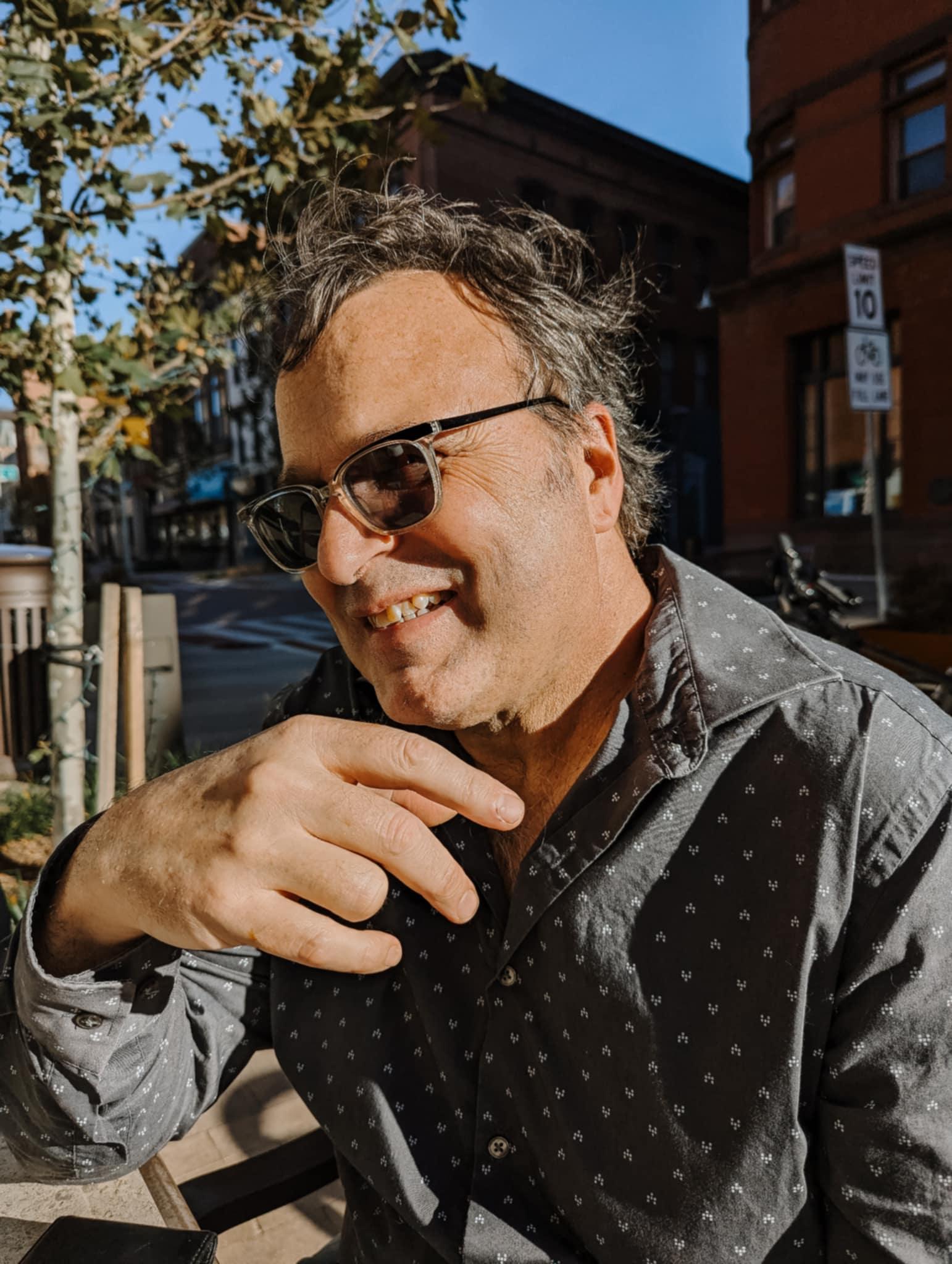 Smiling man with wavy salt-and-pepper hair and dark glasses, wearing grey shirt with white pattern
