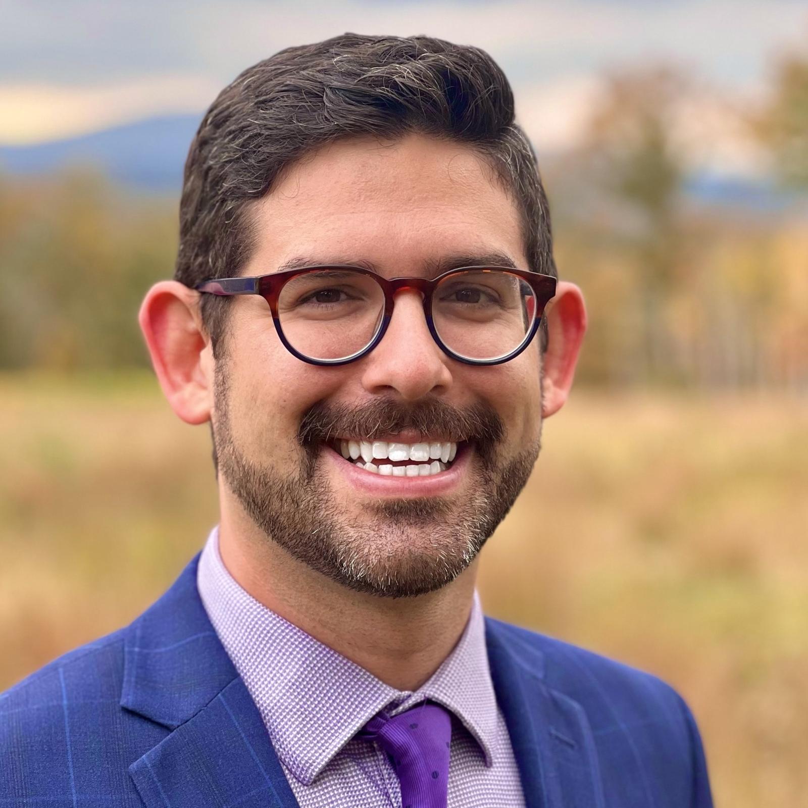 Smiling, bearded man with glasses and dark hair, wearing blue sport coat, purple tie, and lavender shirt.