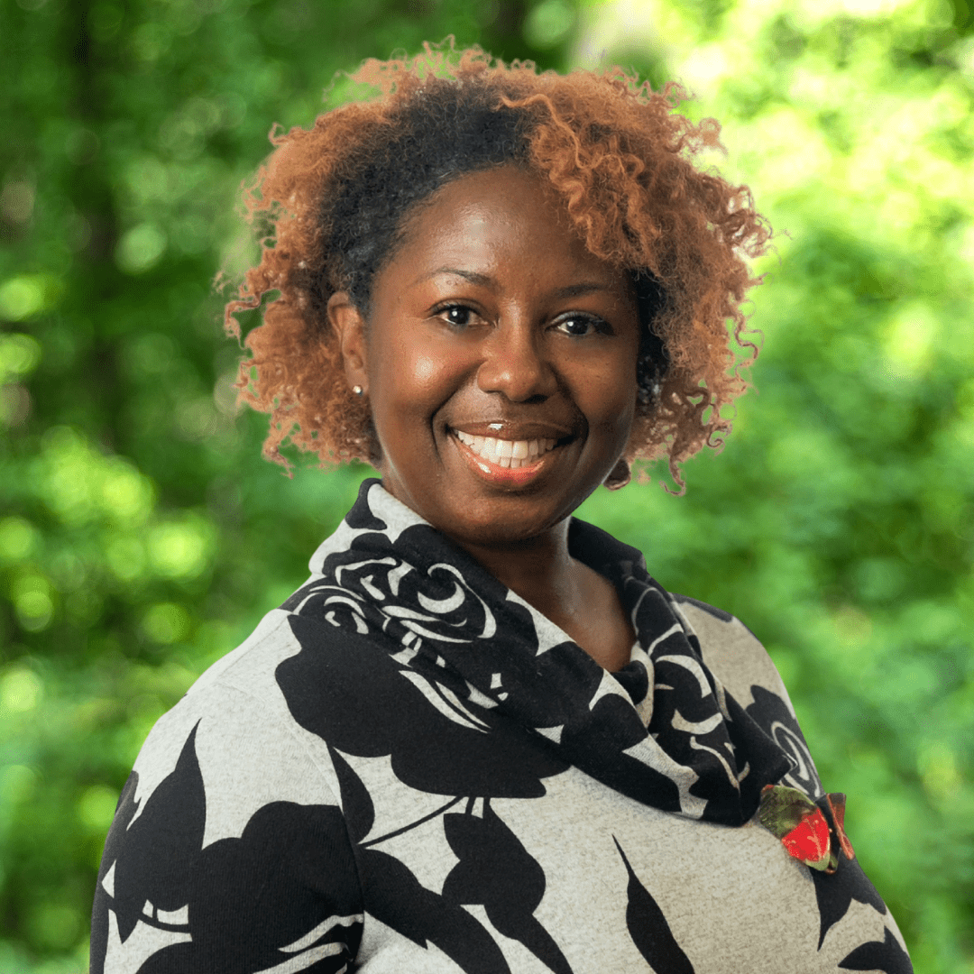Leslie Gillard headshot, women smiling with black and light gray designed sweater, green forest background