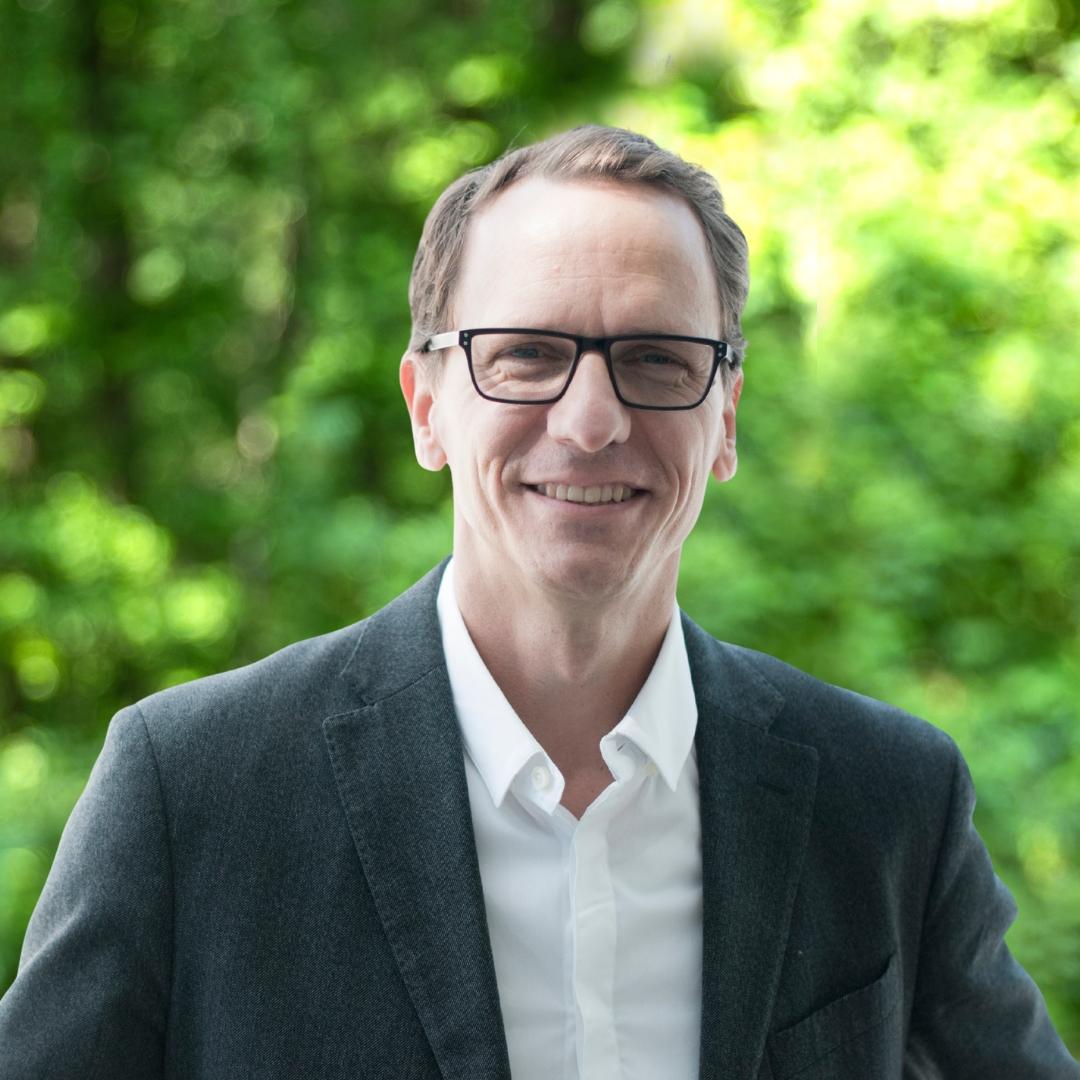 Doug Bothner headshot, man with glass in a dark blue suit jacket smiling directly at camera