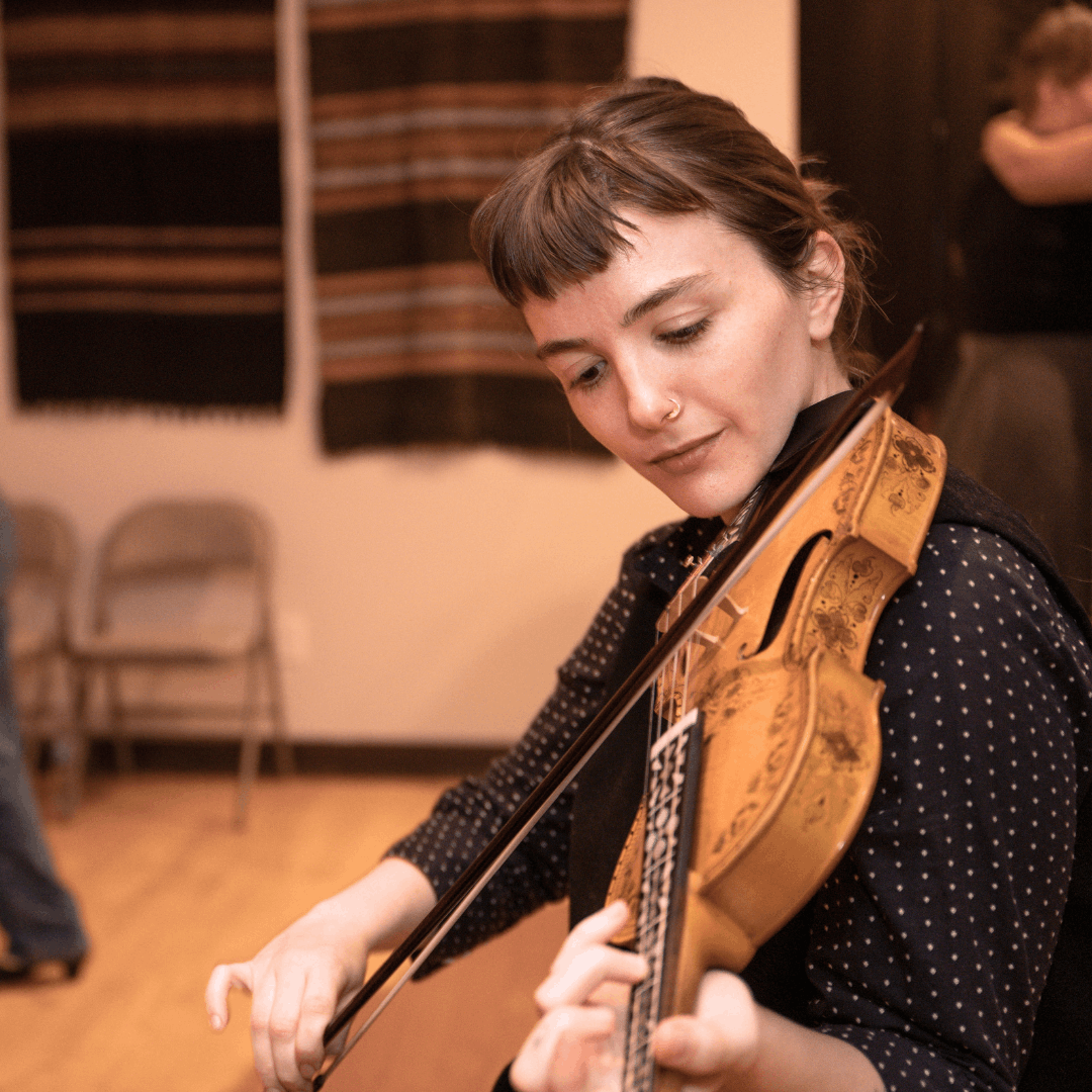 An indoor photograph focuses on a young woman, Georgia Beatty, playing a Norwegian fiddle in the foreground. She is wearing a dark, polka-dot top with a black vest and has short bangs. The instrument has a visible, ornate design on its front. In the blurred background, there are several chairs and what appears to be a patterned textile or tapestry hanging on the back wall.