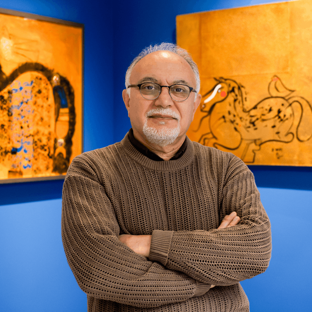 Man posed with cross arms in a brown sweater, in front of corner of room painted blue with a large painting on each wall.