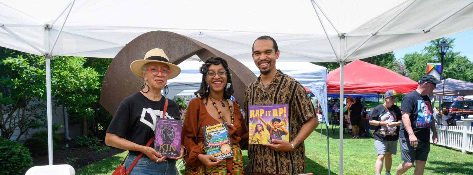 two woman and a man stand posed smiling with a book in each of their hands