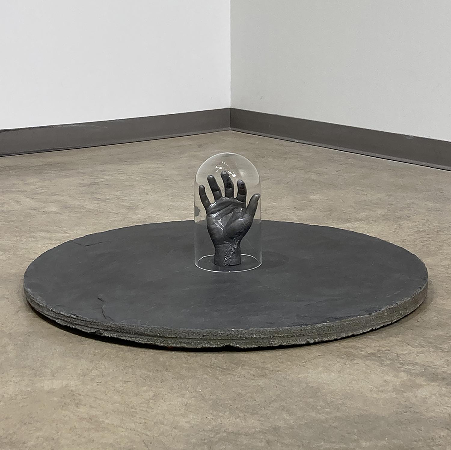 Enclosed in glass on an island of rock, is an empty hand yearning. In an invisible prison of isolation is the unattainable human touch. The granite slab is like a barren desert island upon which the human touch is stranded; barricaded in the glass dome the hand can speak its pain only visually. The work is about the sense of isolation from social distancing.