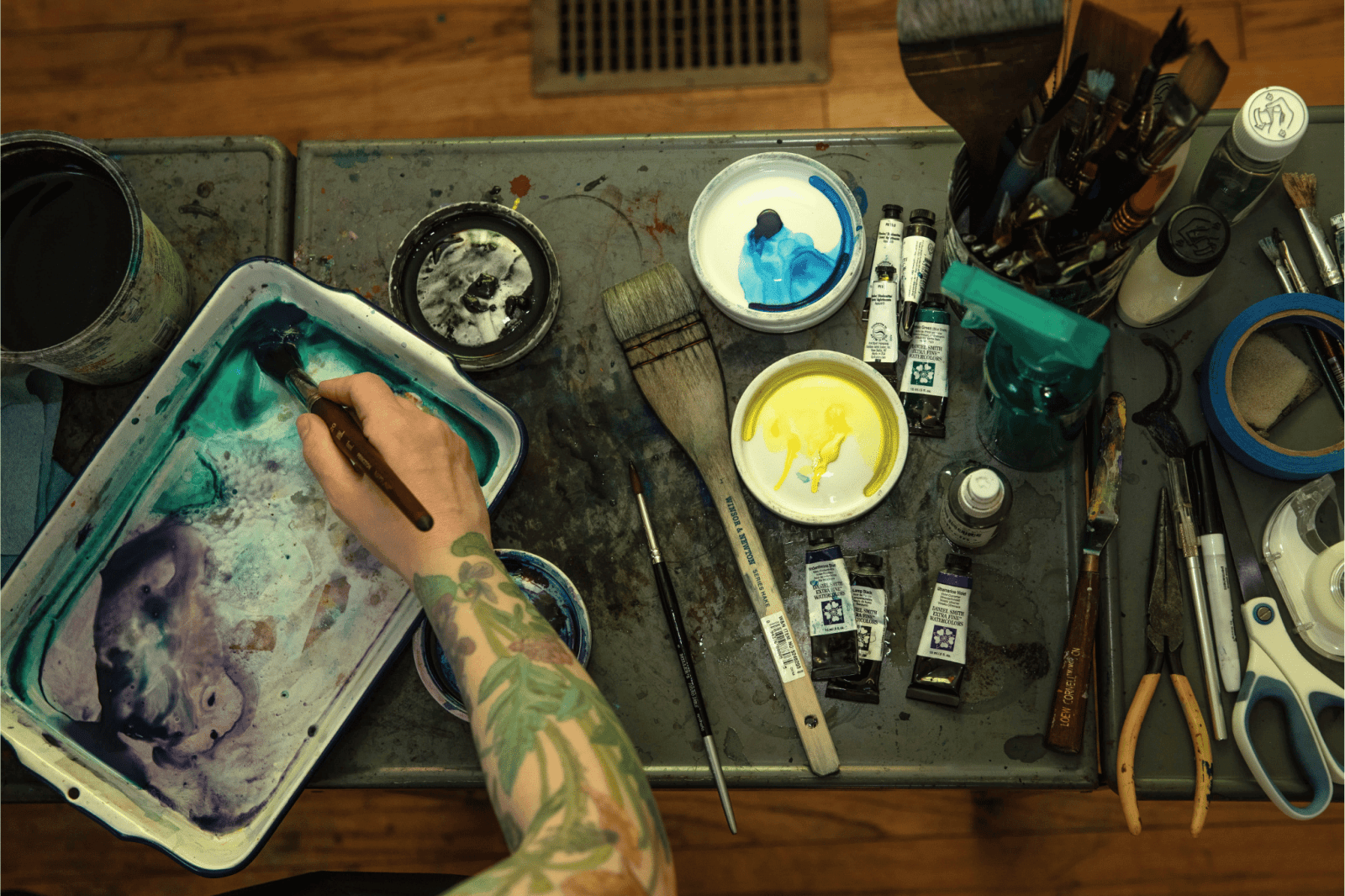 Birdseye view of a tattooed painters' hand mixing paint on a palette surrounded by art supplies