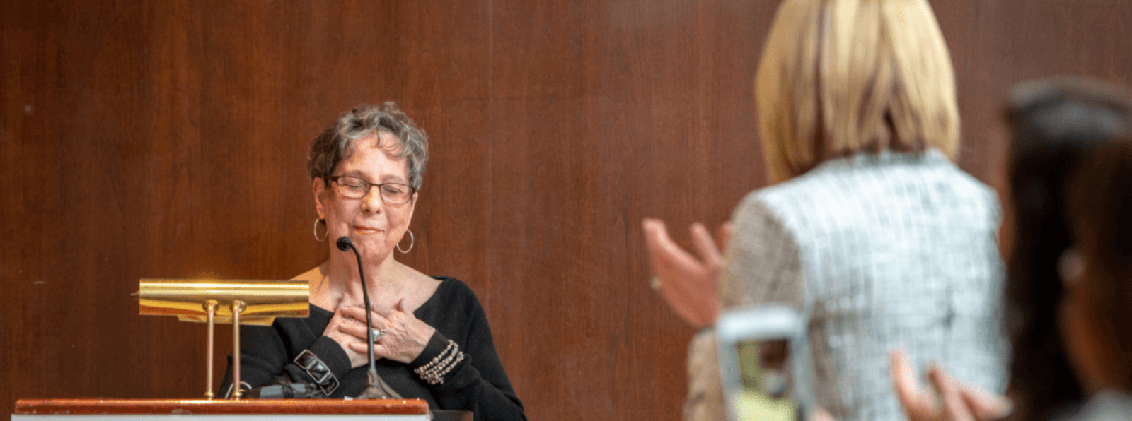 Two people clapping for the Poet Laureate who is standing behind a podium.